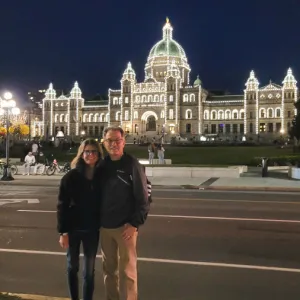 A smiling couple standing in front of the British Columbia Parliament Buildings outlined with glowing white lights
